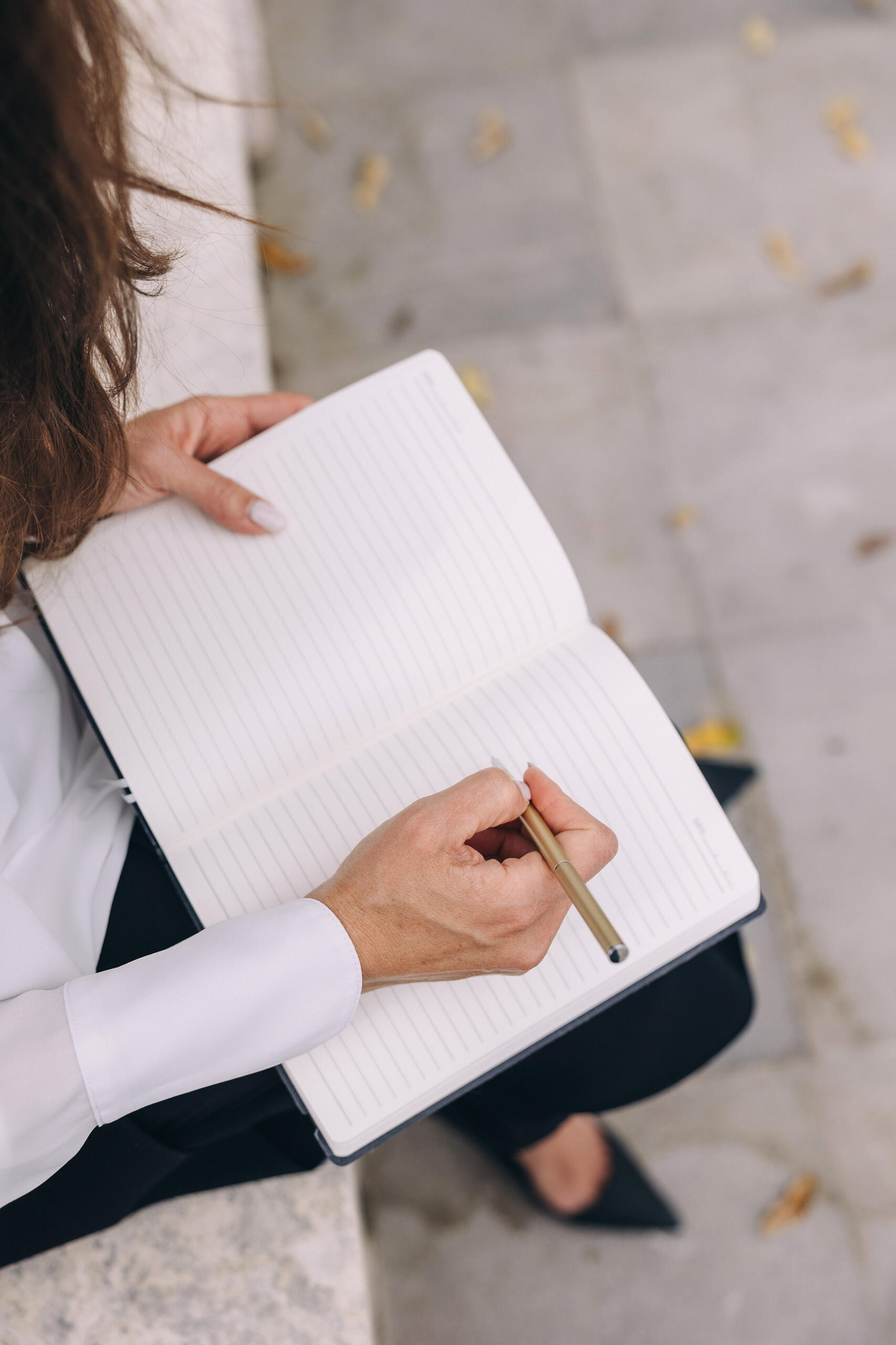 Woman wearing a white shirt and black slacks, sitting outside and writing in a tablet.