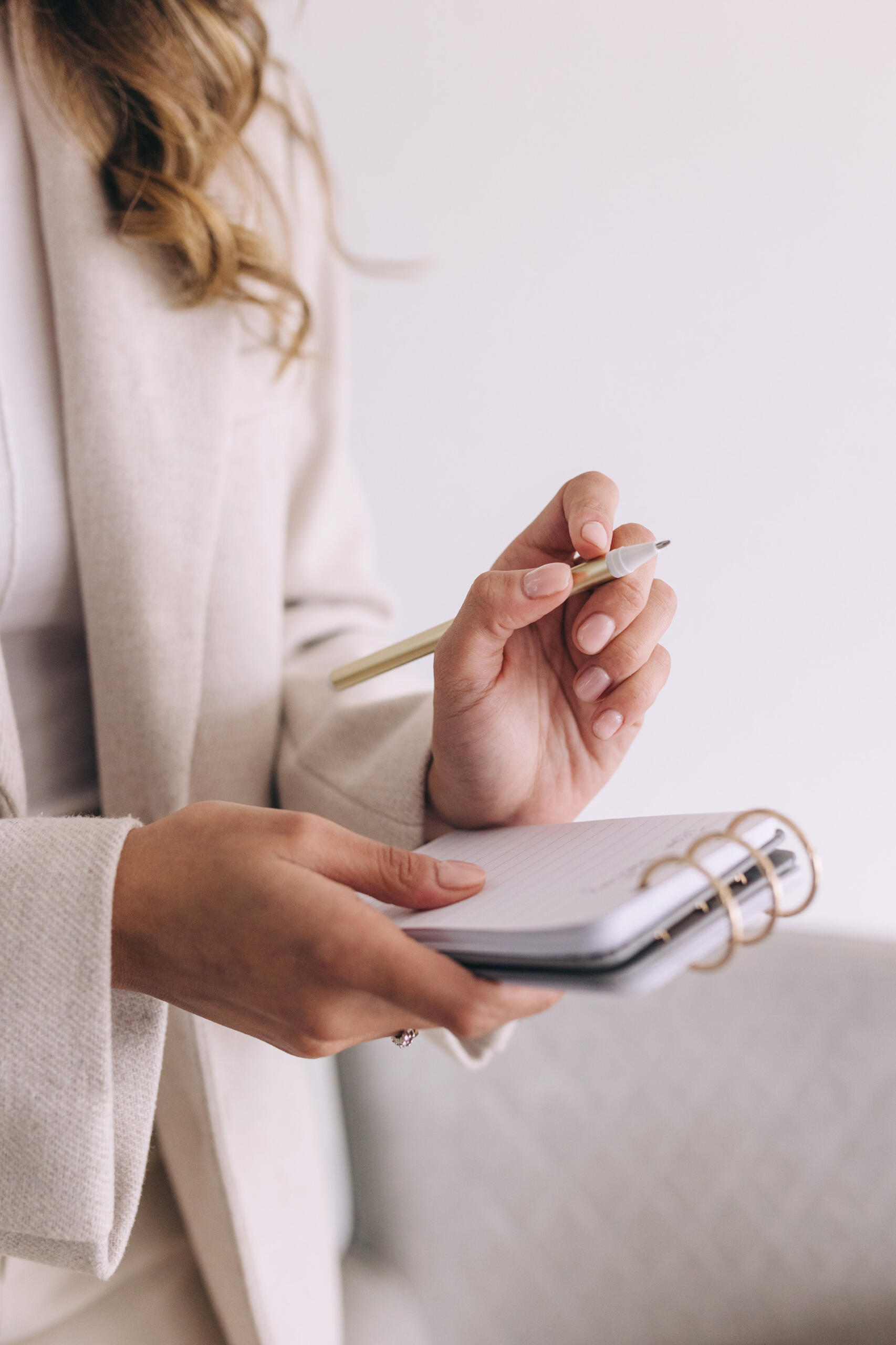 A business woman wearing a cream suit and shite blouse, standing and holding a pen and open notebook.